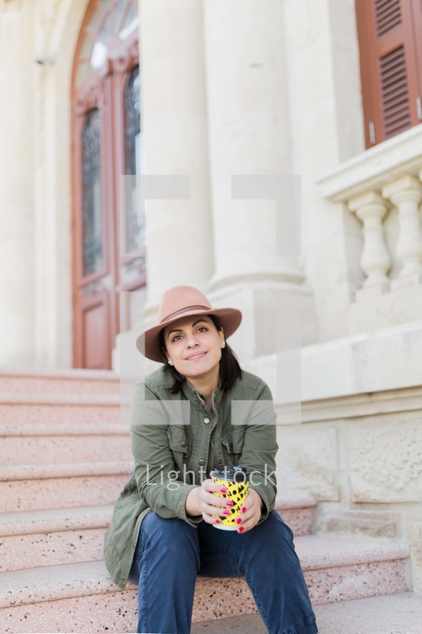 Portrait of a woman sitting on the steps of a building holding a to-go coffee cup.