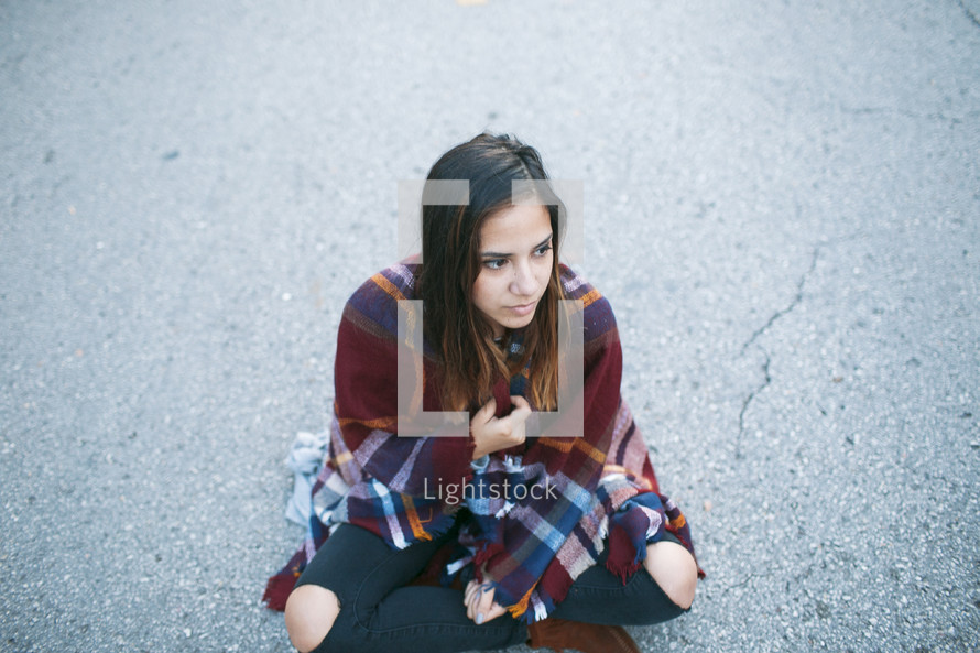 a lost teen wrapped in a blanket sitting on asphalt 
