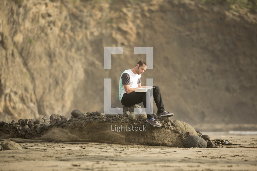 man sitting on rock reading a Bible on a beach 