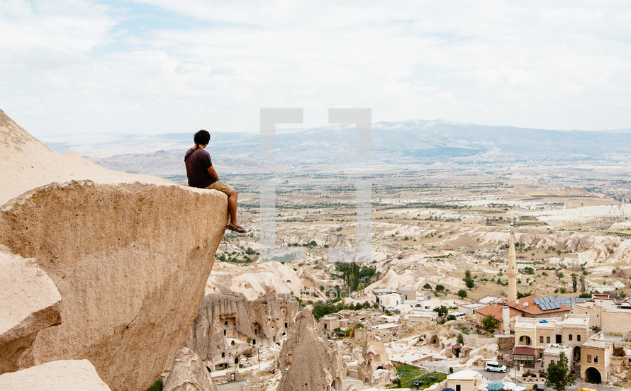 a man sitting at the edge of a cliff looking out at the western town below 
