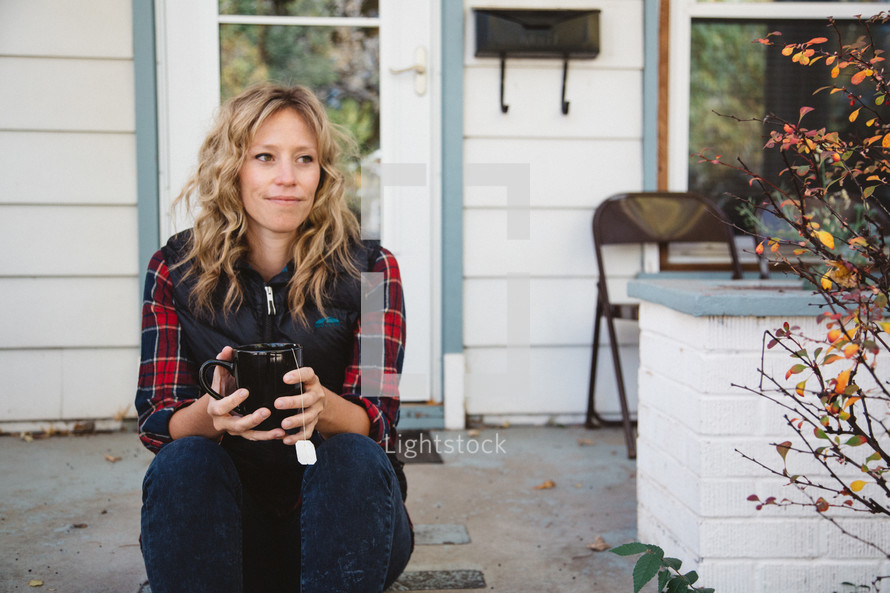 woman sitting on a stoop in front of her house holding a mug of tea 