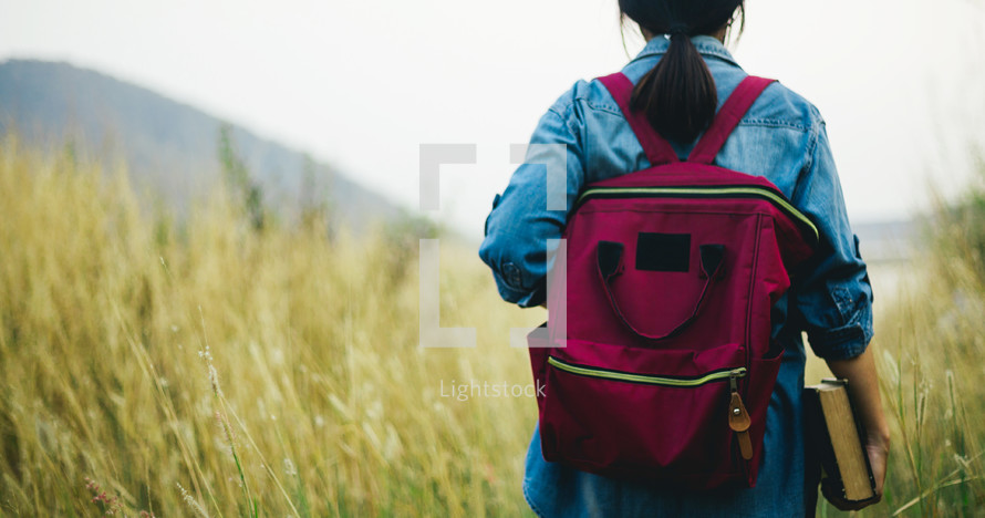 a girl walking through a field carrying a Bible 