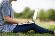 a woman sitting in the grass typing on a laptop 