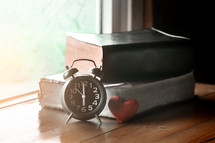stacked Bibles, alarm clock, and red heart in a window 