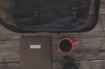 A bag, notebook, coffee mug and french press on wood