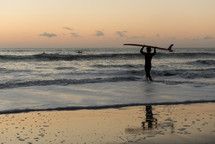 Silhouette of a person holding surf board standing in the ocean at dusk with a reflection n the wet sand on the beach.