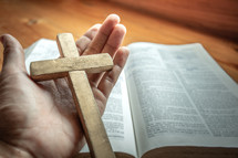 wooden cross in open hands in prayer over a Bible 