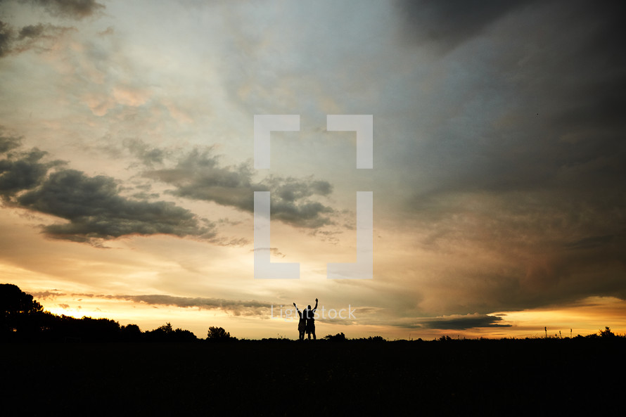 silhouettes of a couple with raised hands under cloudy skies at sunset 
