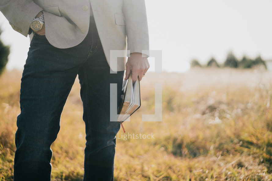 a man holding a Bible at his side outdoors in a field 