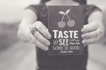woman holding a sign that reads, Taste and see that the lord is good, Psalm 34:8
