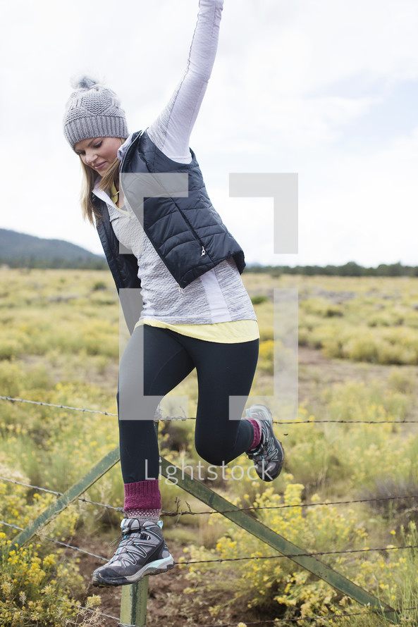A young women climbing over a barbed wire fence in a field of wildflowers.