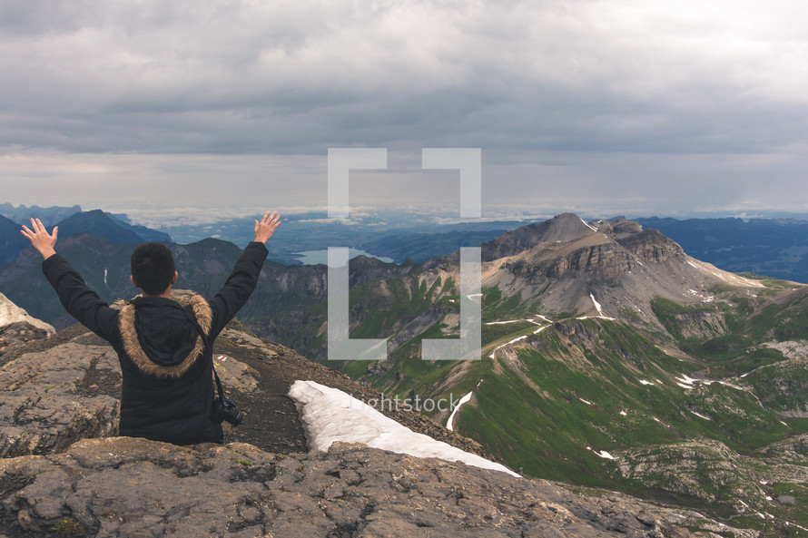 a man with a camera and raised hands sitting on a mountaintop 