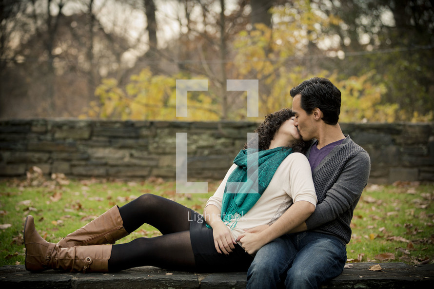 Woman leaning back on on man kissing as they sit in the park.
