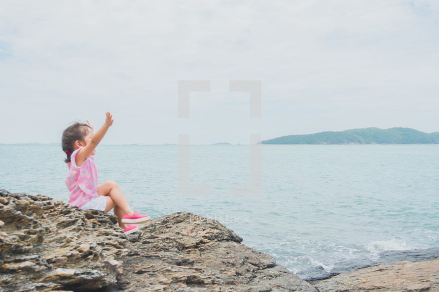 a little girl sitting on a rock on a shore 