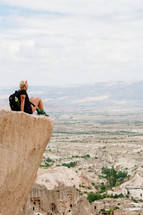 a woman sitting at the edge of a cliff looking out at a western town below 