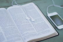 Iphone on a table with earbuds on an open bible.