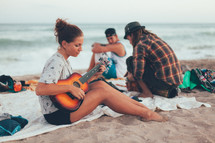 young woman sitting on a beach playing her guitar and friend listening 