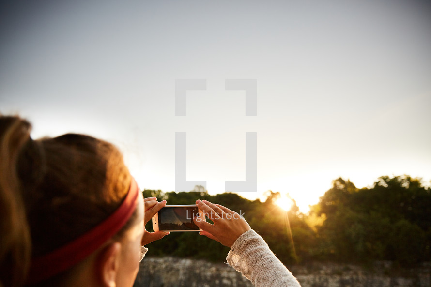 a woman taking a picture of the sunset with her cellphone 