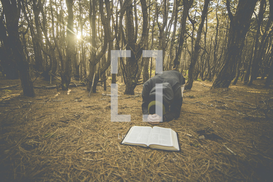 a man kneeling in prayer in front of a Bible in a forest 