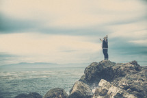 Man praising God on an ocean-side rocky cliff.