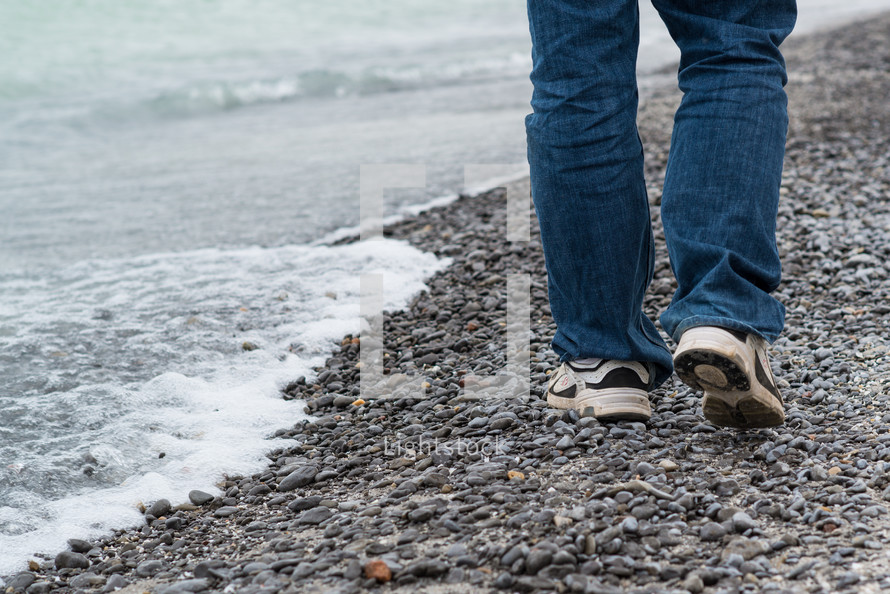 A man's legs walking on a gravel shore.