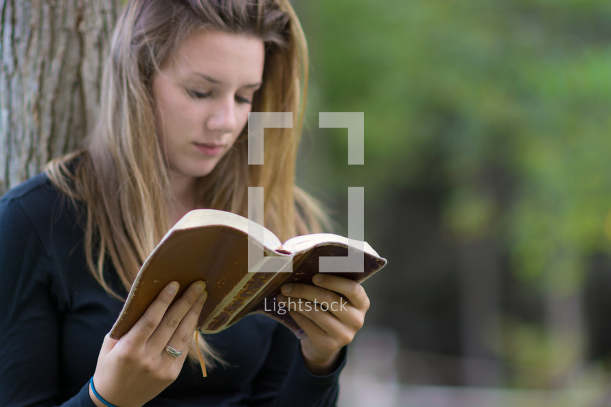 Girl sitting outside, leaning against a tree, reading a bible.