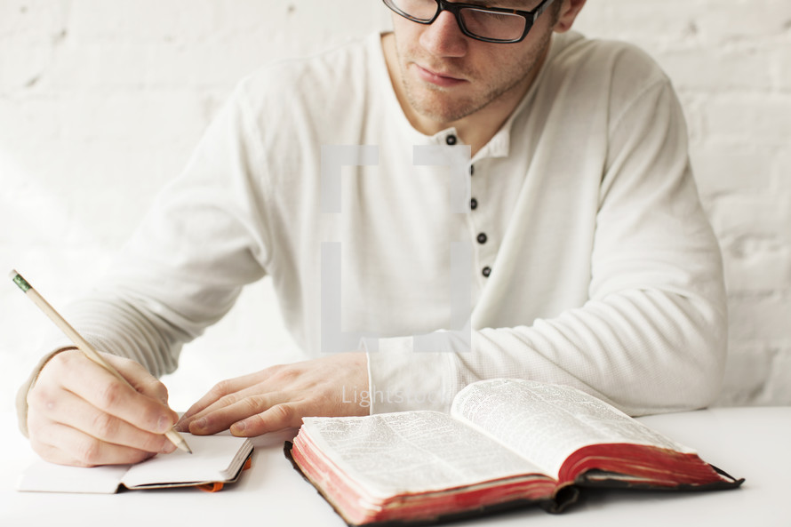Man writing in a journal with a pencil next to an open Bible.