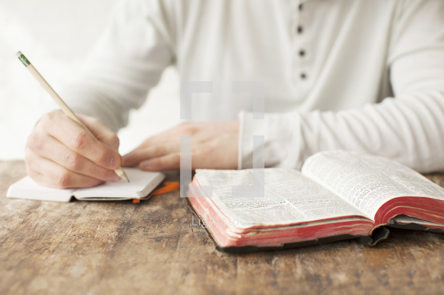 Writing in a journal with a pencil, on a wooden table with an open Bible.