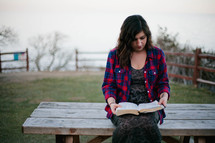 A woman sitting on a bench outdoors reading a Bible. 