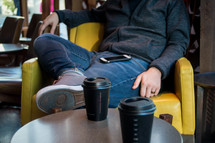 man with a cellphone on his lap sitting in a chair next to a coffee cup 