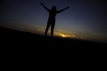 a woman standing with raised arms in worship at sunset 