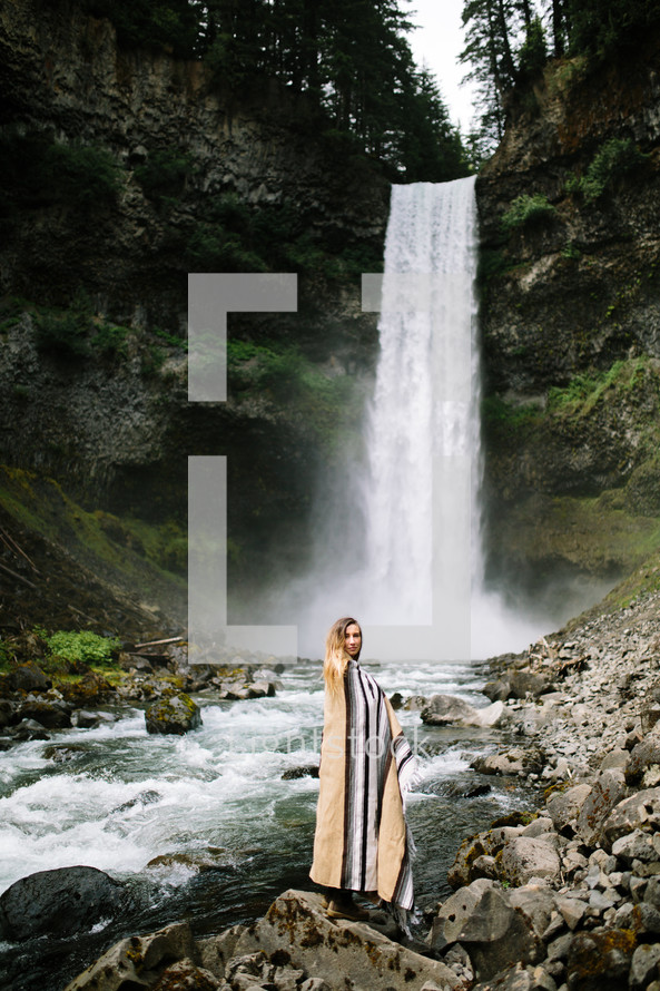 a woman wrapped in a blanket standing in front of a waterfall 