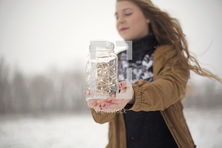 a woman holding a mason jar full of fairy lights outdoors in the snow 
