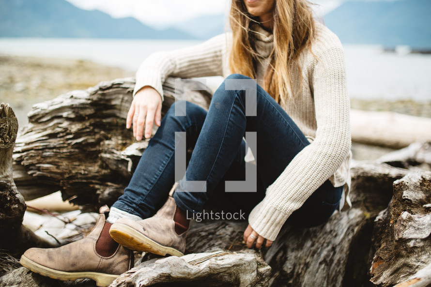 a woman in boots sitting on driftwood on a beach 