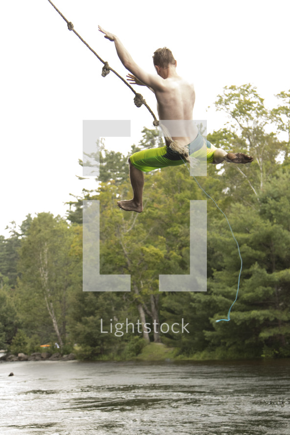 Teenage boy swinging high from rope swing over water and letting go