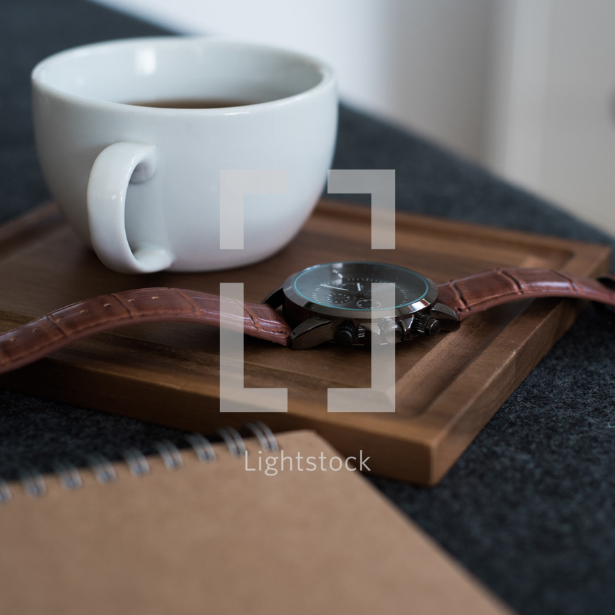 A cup of coffee and a wristwatch on a wooden board next to a spiral notebook.