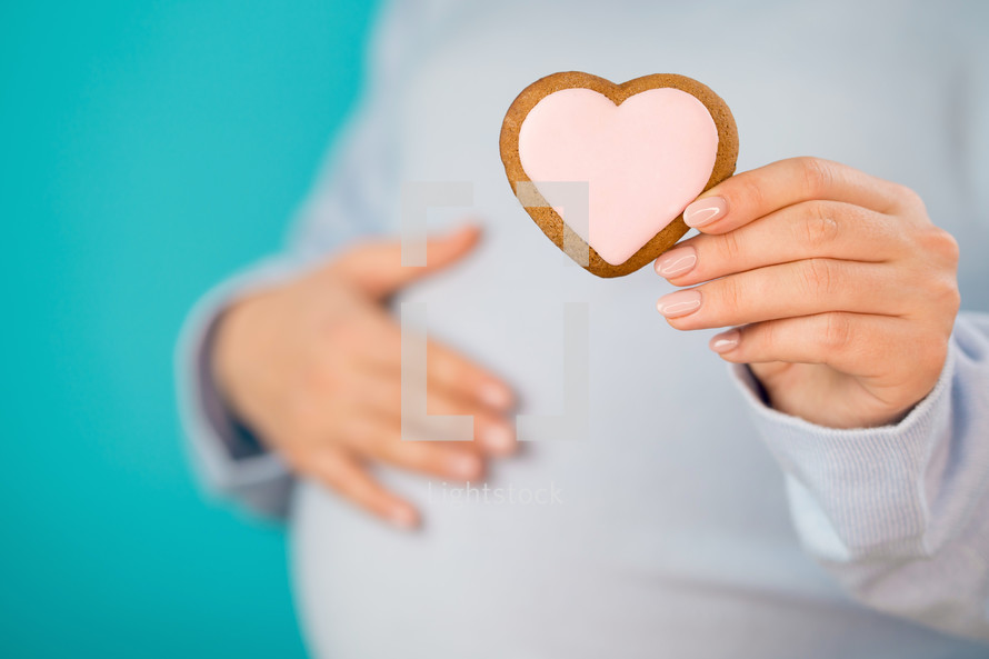 Pregnant woman holding heart-shaped cookie on tummy belly background. Young girl in blue wearing expecting baby. Maternity, motherhood, pregnancy, love concept