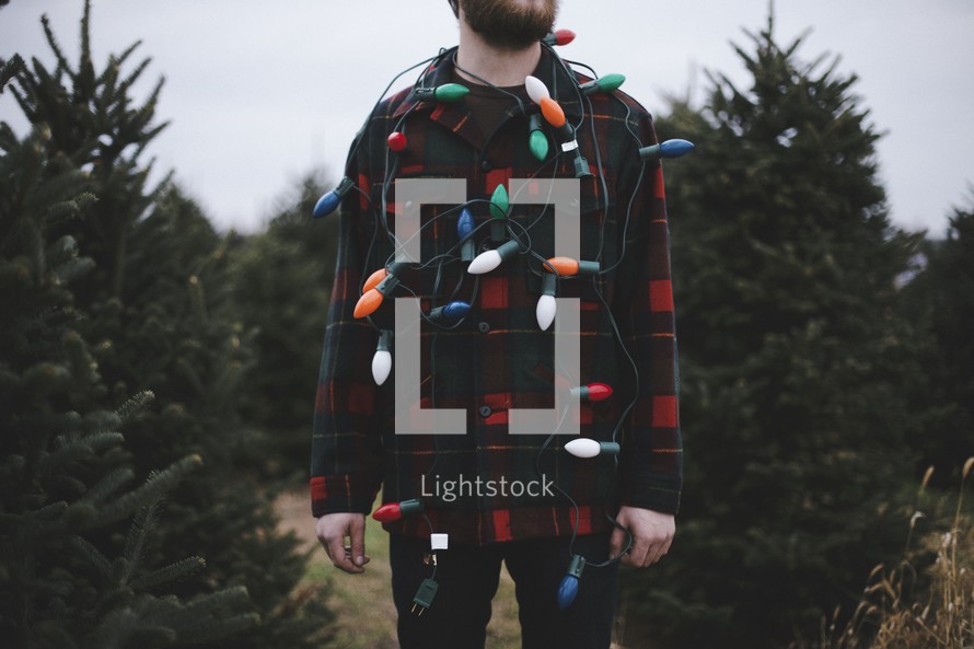 a man in a plaid shirt with Christmas lights standing in a Christmas tree lot 