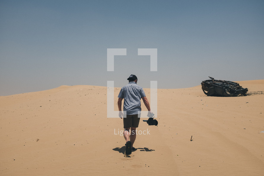a man with a camera walking towards a burned out car in a desert 