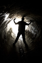 Silhouette of a man straddling water flowing through a sewer drian pipe with arms raised.