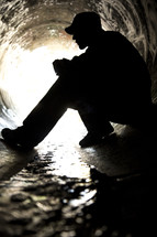 Silhouette of a man sitting in a sewer drian pipe.