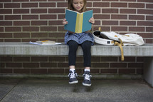 a child sitting on a bench reading while waiting for school 