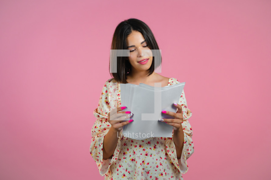 Satisfied woman in floral dress holding files papers isolated on pink background. Pretty girl checks documents, utility bills