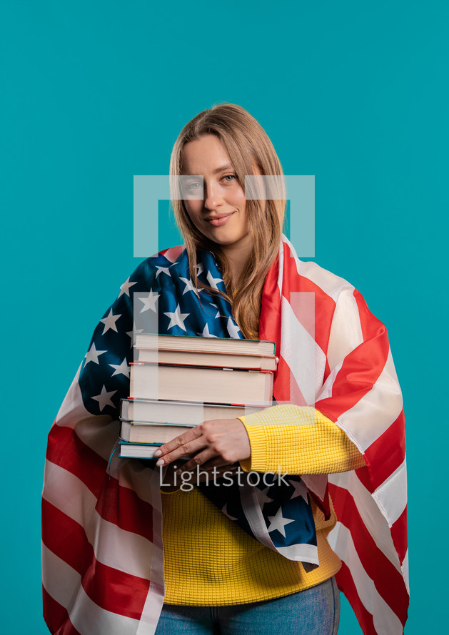 American woman student holds stack of university books from college library on blue background. Happy girl smiles, she is happy to graduate in USA, education abroad concept