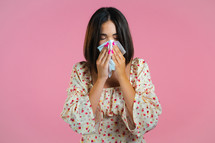 Woman sneezes into tissue. Isolated girl on pink studio background. Lady is sick, has a cold or allergic reaction. Coronavirus, epidemic 2024, illness concept.