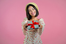 Excited woman holding gift box and gives it by hands to camera on pink wall background. Girl smiling, she is happy with present. Studio portrait.