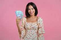 Amazed happy excited woman showing money - U.S. currency dollars banknotes on pink wall. Symbol of success, gain, victory