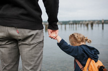 a father and toddler girl holding hands on a river shore 