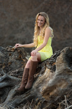 A woman in boots and a yellow dress sitting on a rock outdoors. 