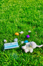 face mask, stuffed animal, and Easter eggs scattered in the grass 